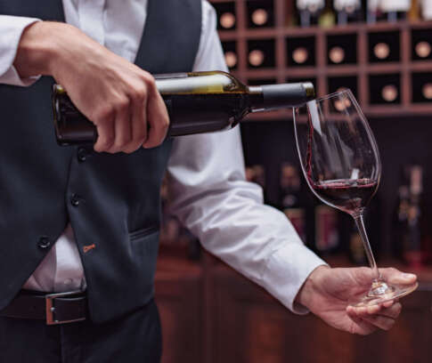 Cropped view sommelier pouring red wine from bottle into glass at table in cellar